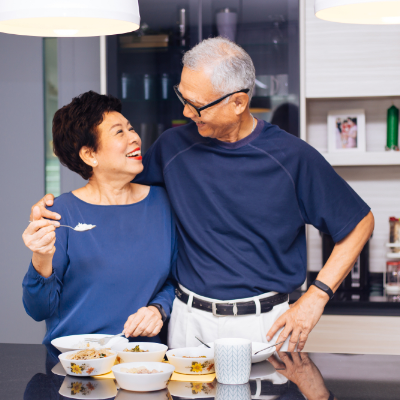 An elderly couple smiling at each other. There is food laid out on a table in front of them and the woman is holding a spoon of food